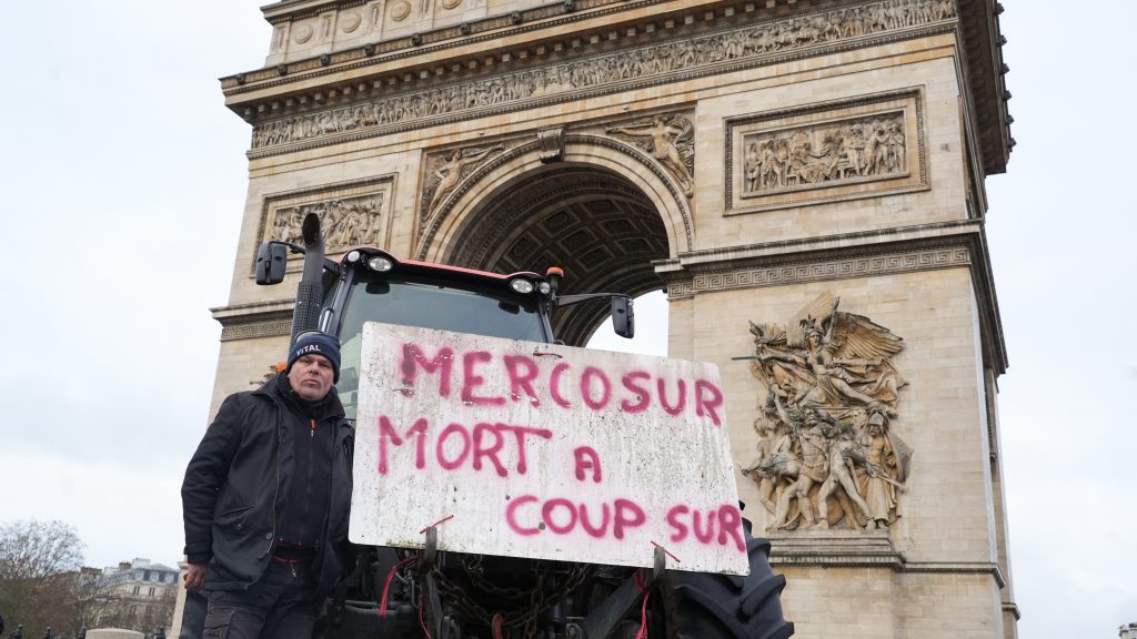 Un homme se tenant devant un tracteur avec une pancarte portant la mention 'MERCOSUR MORT A COUP SUR' devant l'Arc de Triomphe à Paris.